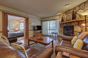 A spacious living room with a stone fireplace, leather sofas, a television, and a large window. - Atrium 104 Downtown Condos with on-site Hot Tub (Breckenridge)