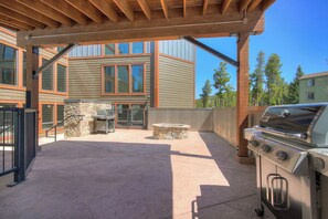 An outdoor patio with a grill and stone seating, wooden pergola, and a view of a multi-story building.