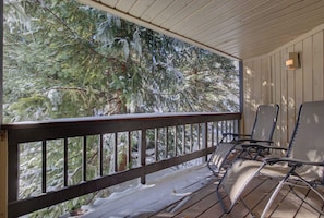 A covered outdoor area with a wooden railing, two lounge chairs, and a view of snow-covered trees. - Atrium 104 Downtown Condos with on-site Hot Tub (Breckenridge)