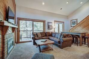 A cozy living room with a sectional sofa, a coffee table, and a fireplace. There are large windows and a wooden staircase. - Lance's West 6 Condo with Pool & Hot Tub (Breckenridge)