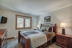Un dormitorio con cama de madera, una mesita de noche con lámpara, un aparador, un televisor montado en la pared y una ventana con vista a árboles.