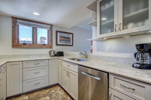A kitchen with white cabinets, a stainless steel dishwasher, a coffee maker, and a window with a view of a snowy landscape. - Lance's West 6 Condo with Pool & Hot Tub (Breckenridge)