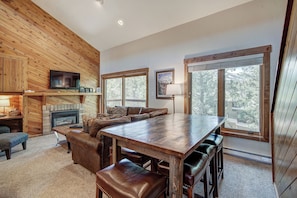 A cozy cabin interior with a wooden dining table, leather seating, a fireplace, and large windows offering a view of the surrounding forest.