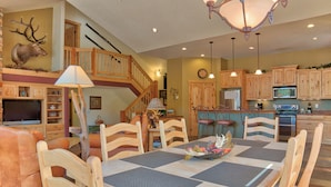 A dining area with a wooden table and chairs, a chandelier, and a kitchen in the background. - Moose Meadow Retreat Private Home with Hot Tub (Breckenridge)