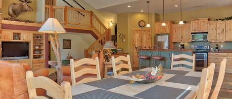 A dining area with a wooden table and chairs, a chandelier, and a kitchen in the background.
