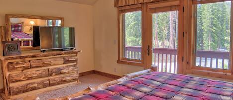 Un dormitorio con cómoda de madera, televisor, cama grande con cubrecama a cuadros y balcón con vistas a árboles.