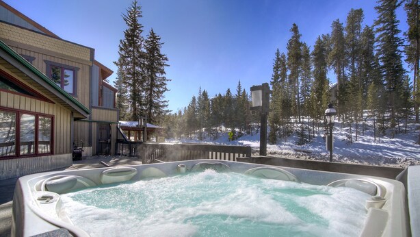 A hot tub surrounded by snow-covered ground and tall pine trees, with a building in the background. - Ten Mile Suites 103 Downtown Condos with On-site Hot Tub (Breckenridge)