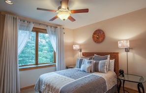 A bedroom with a bed, a ceiling fan, a window with curtains, a wall clock, and two bedside lamps.
