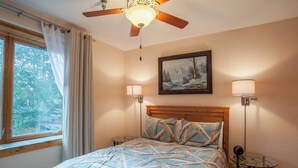 A bedroom with a bed, a ceiling fan, a window with curtains, a painting on the wall, and two bedside lamps. - Ten Mile Suites 103 Downtown Condos with On-site Hot Tub (Breckenridge)