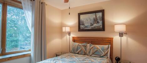 A bedroom with a bed, a ceiling fan, a window with curtains, a painting on the wall, and two bedside lamps.