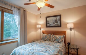 Une chambre à coucher comprenant un lit, un ventilateur de plafond, une fenêtre avec des rideaux, un tableau au mur et deux lampes de chevet.