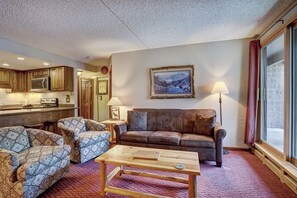 A cozy living room with a brown leather sofa, patterned armchairs, and a wooden coffee table. A painting hangs on the wall, and there is a window with curtains. - Trail's End 201 Downtown Condos with on-site Hot Tub (Breckenridge)