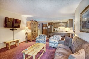 Un salon comprenant un canapé, deux fauteuils, une table basse en bois, un téléviseur, un meuble et un coin cuisine avec un réfrigérateur et un évier.