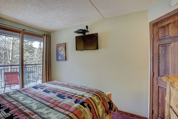 Une chambre avec une grande fenêtre, un lit recouvert d’une couverture aux couleurs vives, un tableau accroché au mur et une porte en bois.