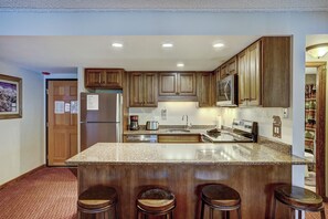 A kitchen with wooden cabinets, a stainless steel refrigerator, a granite countertop, and a built-in oven. There are three stools in front of the counter.