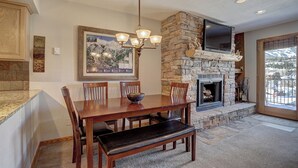 A dining area with a stone fireplace, a television mounted above it, and a view of a snowy landscape through a large window. - Wellington #3 (Townhome with Private Hot Tub) (Breckenridge)