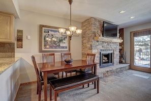 A dining area with a stone fireplace, a television mounted above it, and a view of a snowy landscape through a large window. - Wellington #3 (Townhome with Private Hot Tub) (Breckenridge)