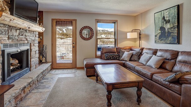 A cozy living room with a stone fireplace, leather sofa, and a large window offering a mountain view. - Wellington #3 (Townhome with Private Hot Tub) (Breckenridge)