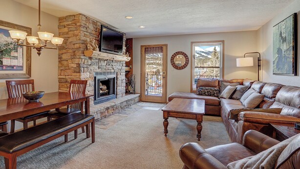 A cozy living room with a stone fireplace, leather sofa, wooden dining table, and a television mounted above the fireplace. - Wellington #3 (Townhome with Private Hot Tub) (Breckenridge)