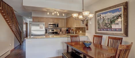 Una cocina con armarios de madera, electrodomésticos de acero inoxidable, una zona de comedor con una mesa y sillas de madera, y una escalera a la izquierda.
