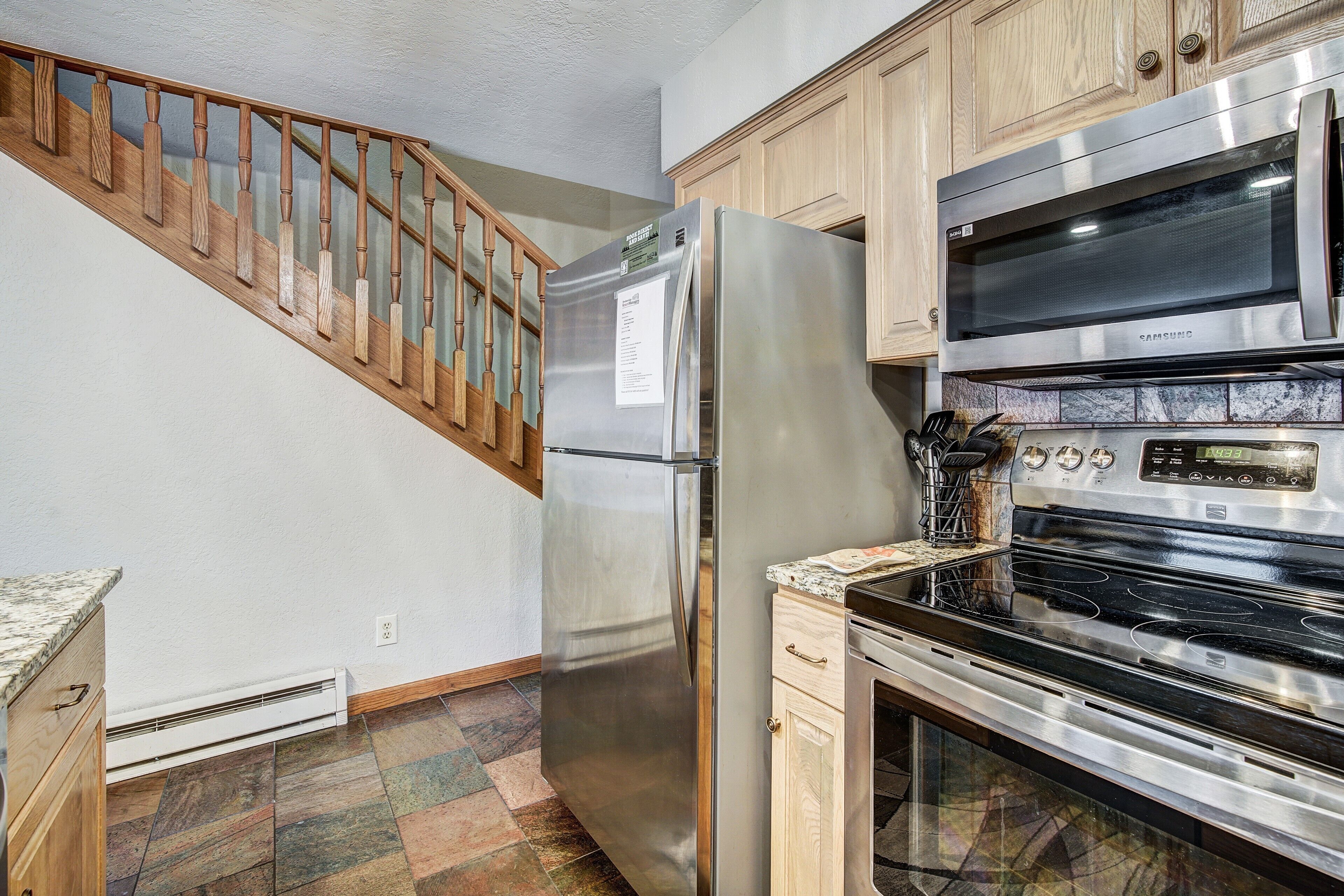 A kitchen with a stainless steel refrigerator, a microwave, and an oven. There is a wooden staircase with a glass railing leading to another level.