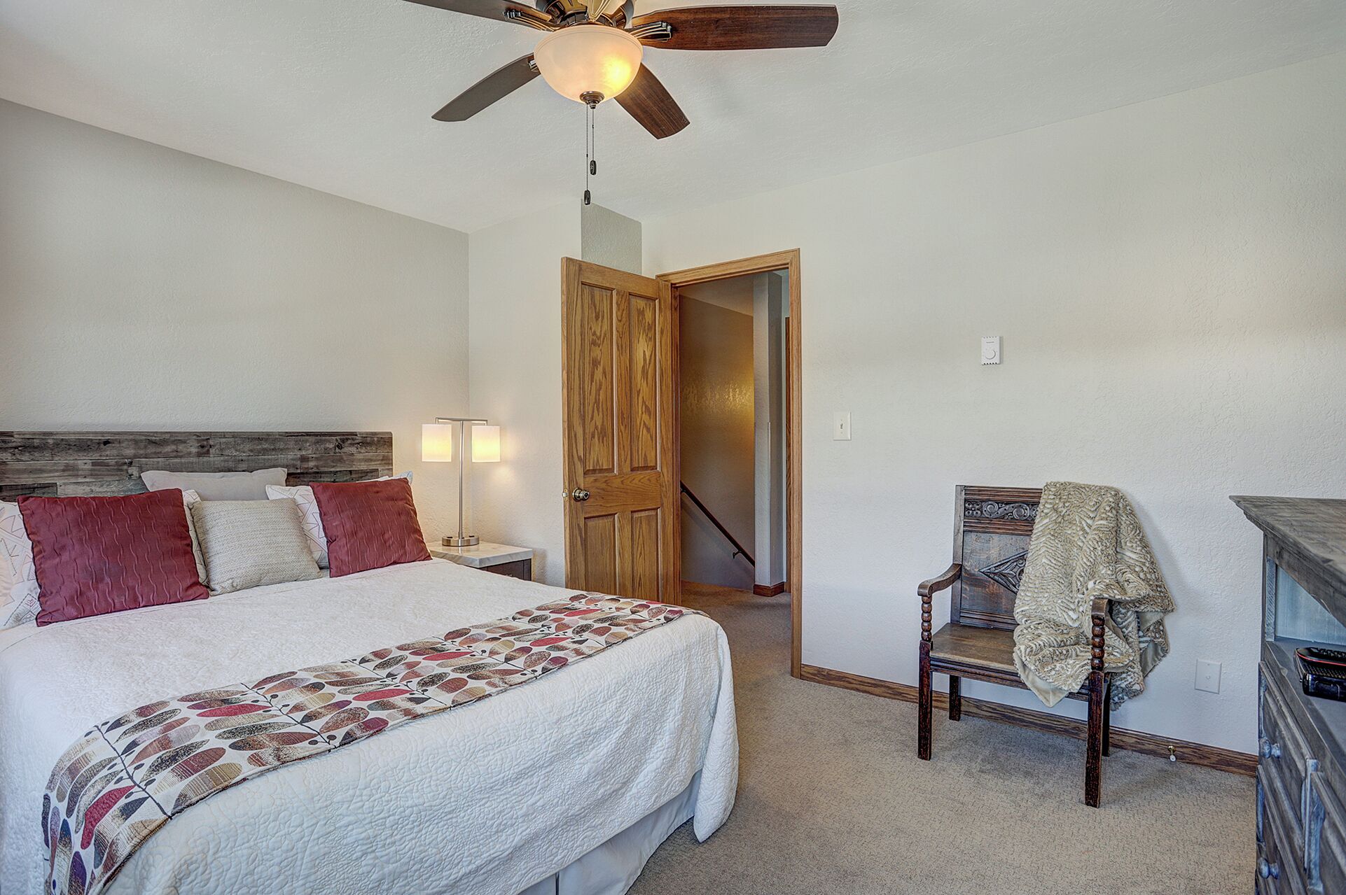 A bedroom with a bed, a wooden headboard, a ceiling fan, a nightstand with a lamp, a wooden chair, and a dresser.