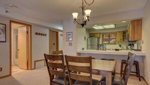 A dining area with a wooden table and chairs, a chandelier, and a kitchen in the background. - Atrium 207 Downtown Condos with on-site Hot Tub (Breckenridge)