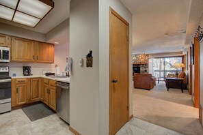 A kitchen with wooden cabinets, a microwave, and a coffee maker. There is a door leading to a living room with a couch and a stone fireplace.