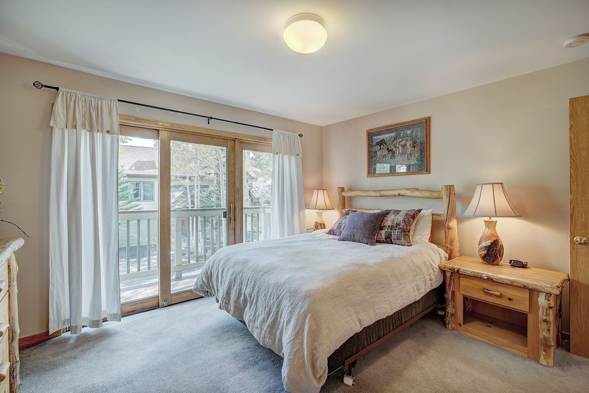 A bedroom with a large bed, two bedside tables with lamps, a wooden headboard, a painting on the wall, and a sliding glass door leading to a balcony.