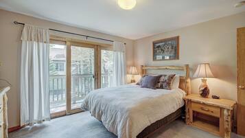A bedroom with a large bed, two bedside tables with lamps, a wooden headboard, a painting on the wall, and a sliding glass door leading to a balcony.