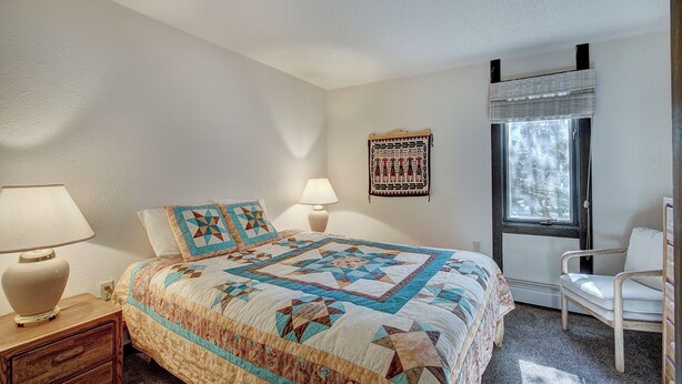 A bedroom with a bed, two bedside lamps, a chair, a window with blinds, and a wall hanging. - Gold Camp II E87 Condo (Breckenridge)