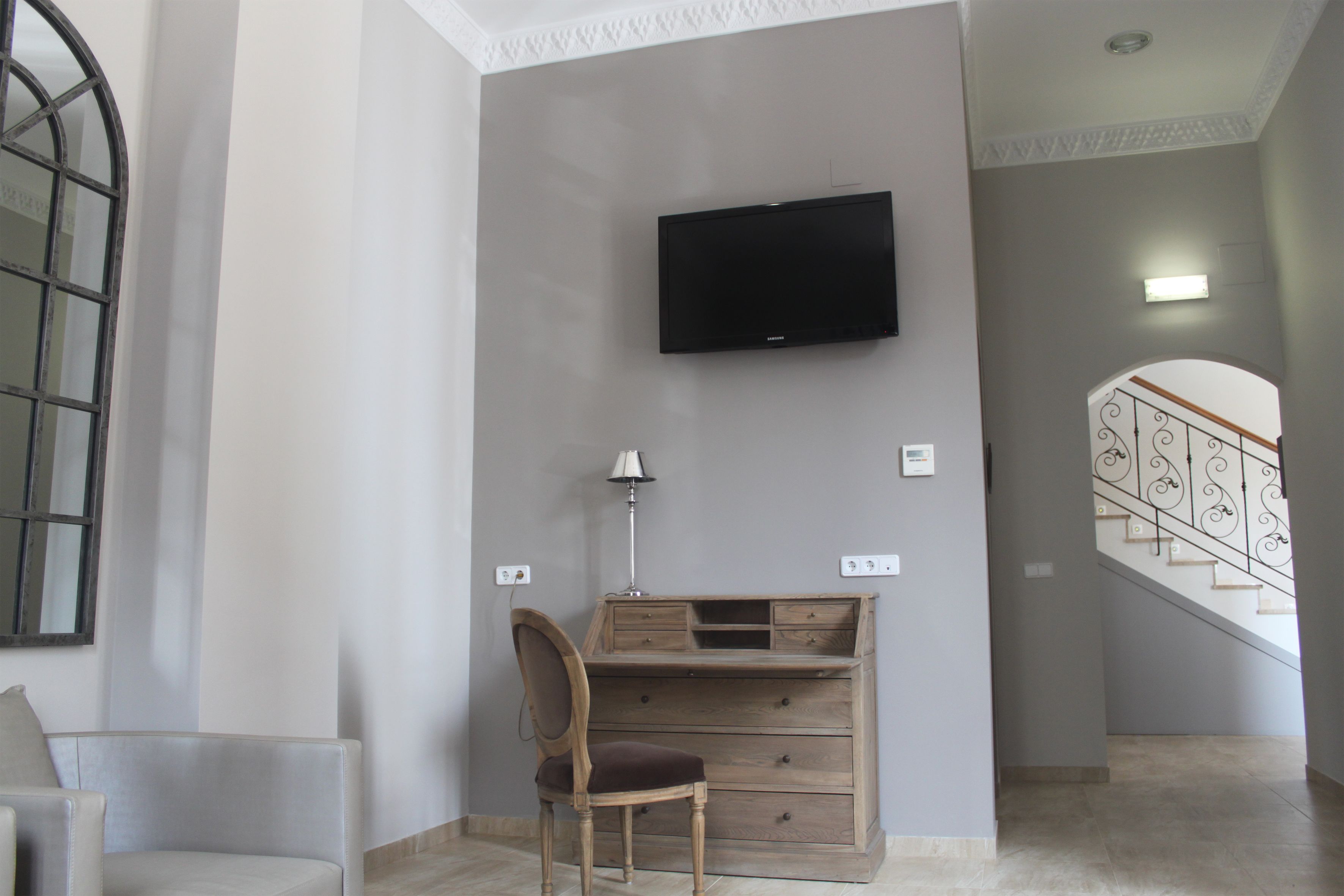 A room with a flat-screen TV mounted on the wall, a wooden desk with drawers, a chair, and a lamp.