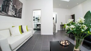 Un moderno salón con un sofá blanco, una mesa de centro de vidrio y un jarrón con flores. Hay un cuadro en la pared y se ve a otra habitación.