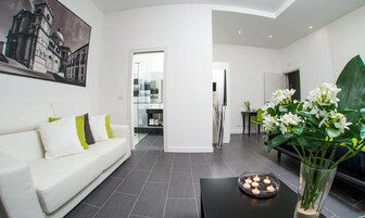 Un moderno salón con un sofá blanco, una mesa de centro de vidrio y un jarrón con flores. Hay un cuadro en la pared y se ve a otra habitación.
