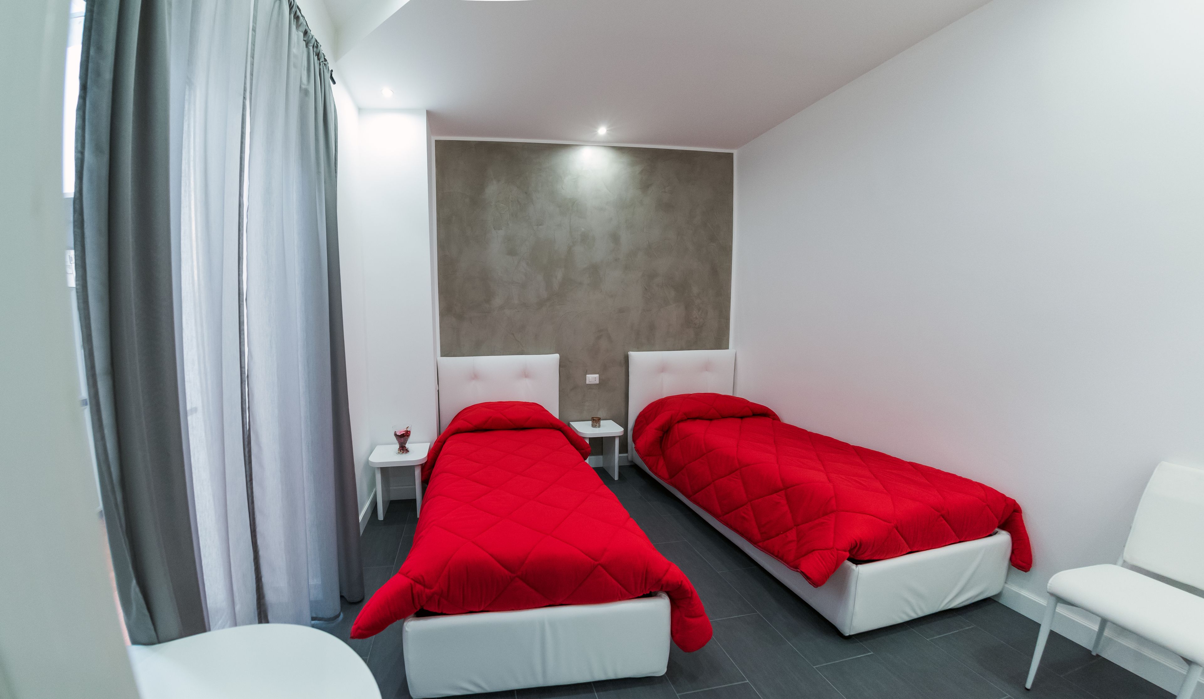 A modern hotel room with two single beds, a small bedside table, a chair, and a large window with curtains.
