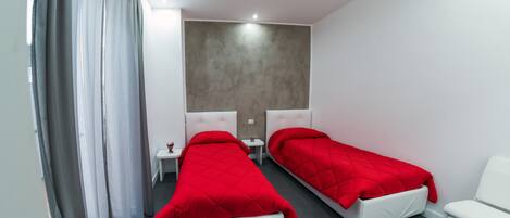 A modern hotel room with two single beds, a small bedside table, a chair, and a large window with curtains.