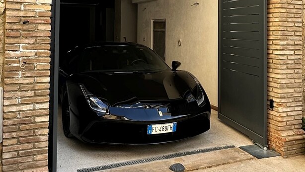 Un coche deportivo negro estacionado en un garaje con fachada de ladrillo y una puerta metálica.