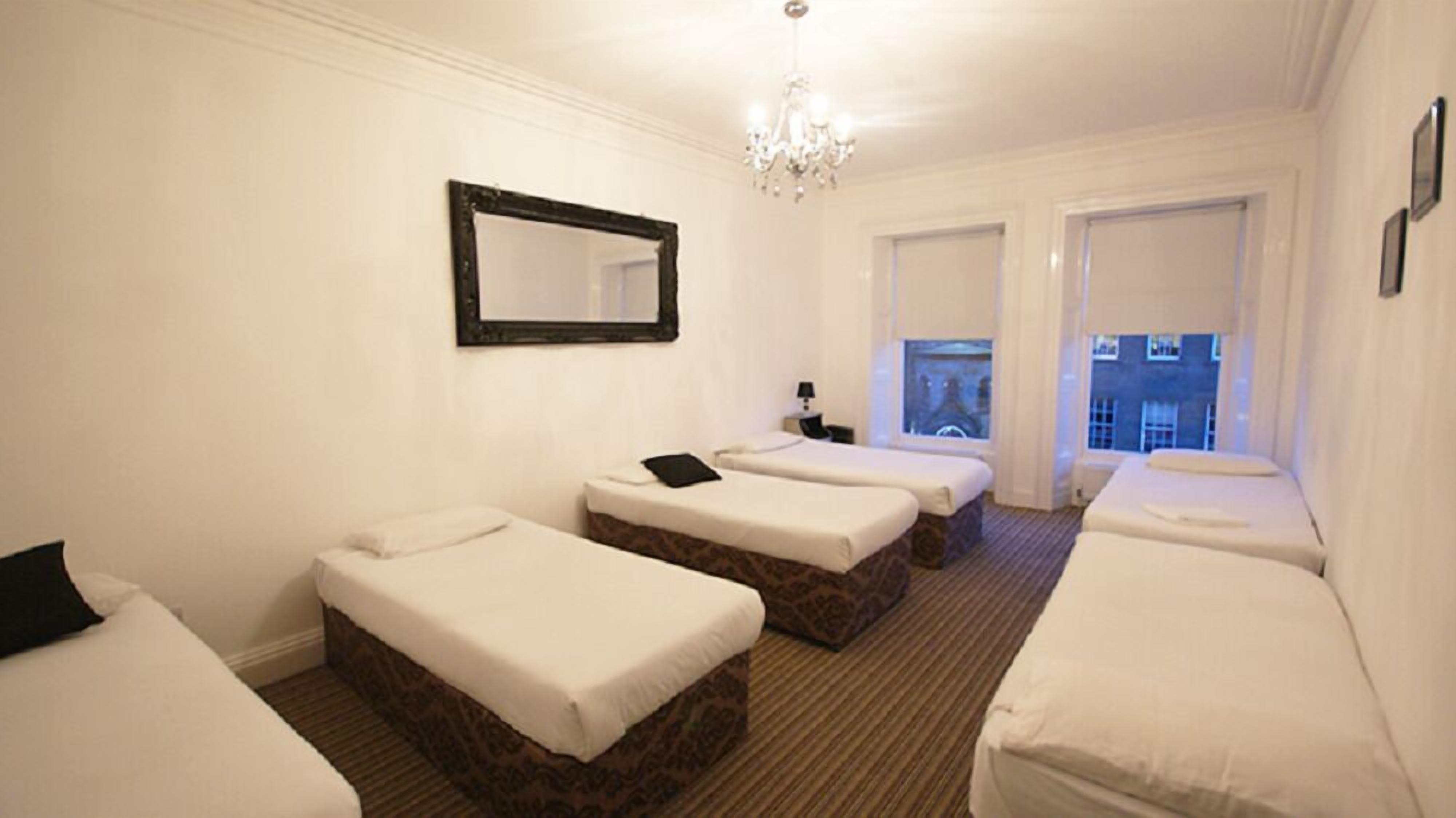 A hotel room with three single beds, a large mirror, a chandelier, and a view of the city through the windows.