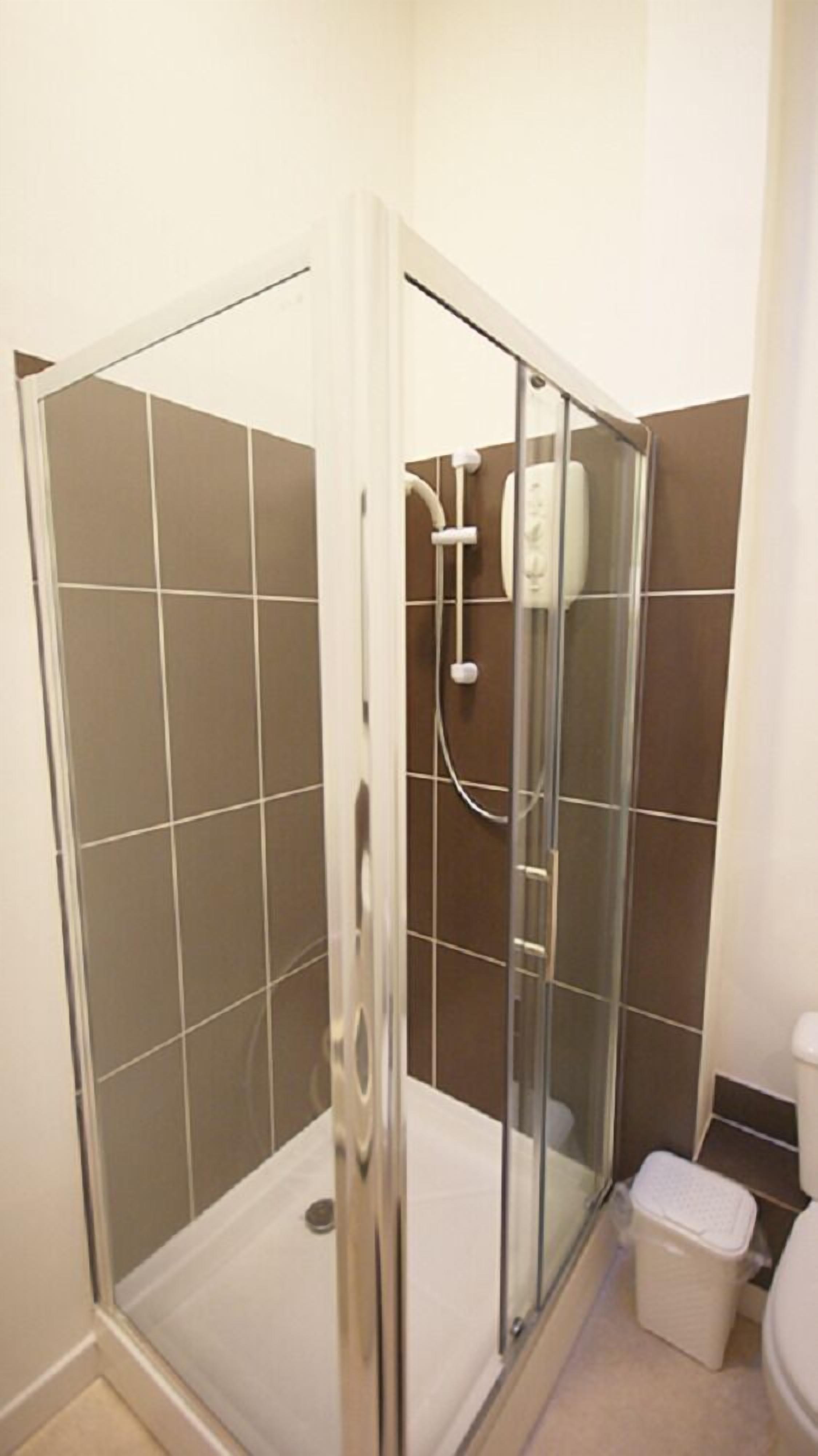 A glass-enclosed shower stall with a white toilet and a trash bin in a bathroom with brown tiles.
