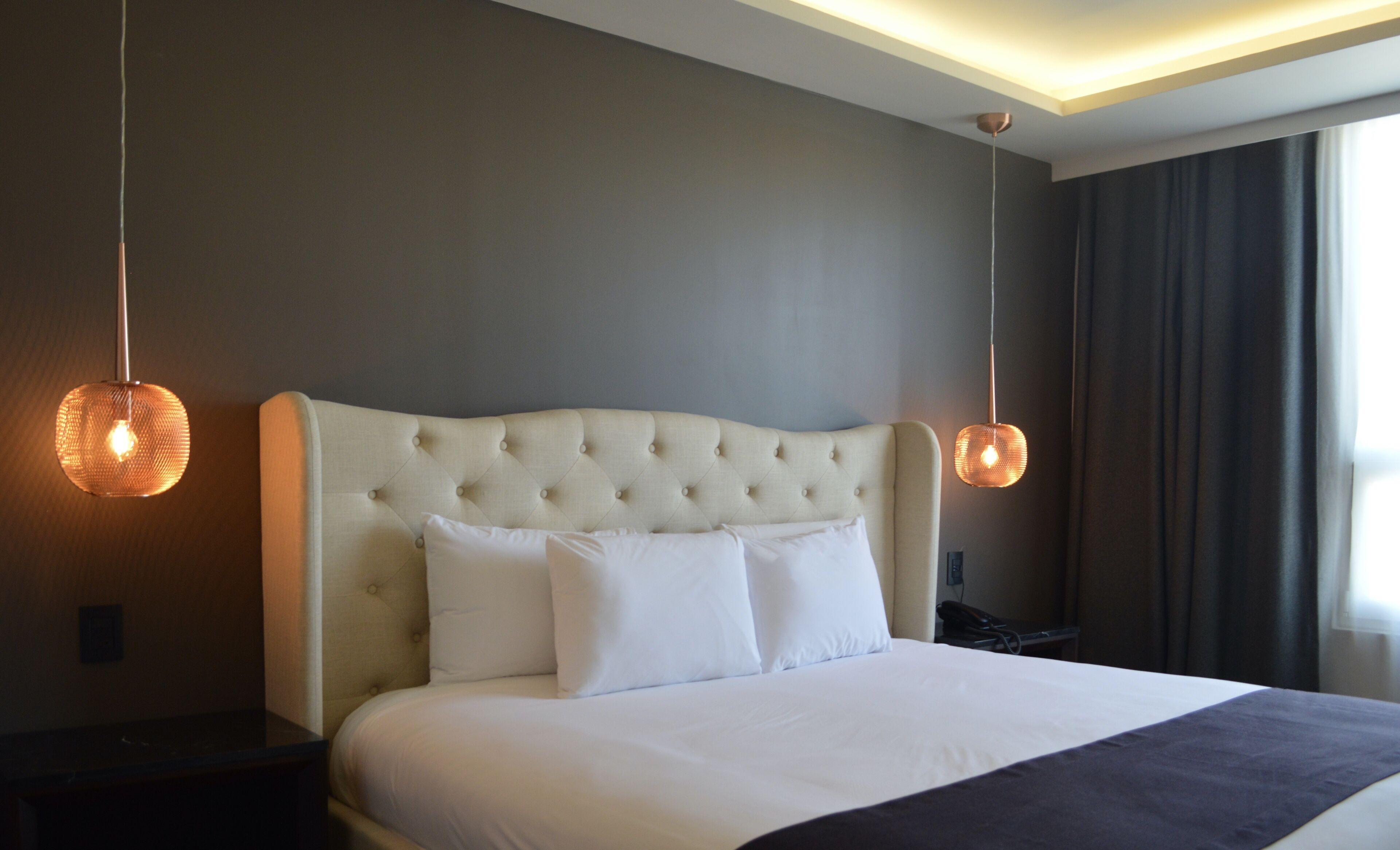 A neatly made bed with a tufted headboard, two bedside lamps, and a dark curtain to the right.