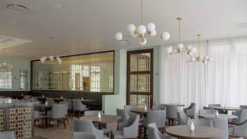 A restaurant with round tables and grey chairs, a decorative ceiling, and large windows with curtains.