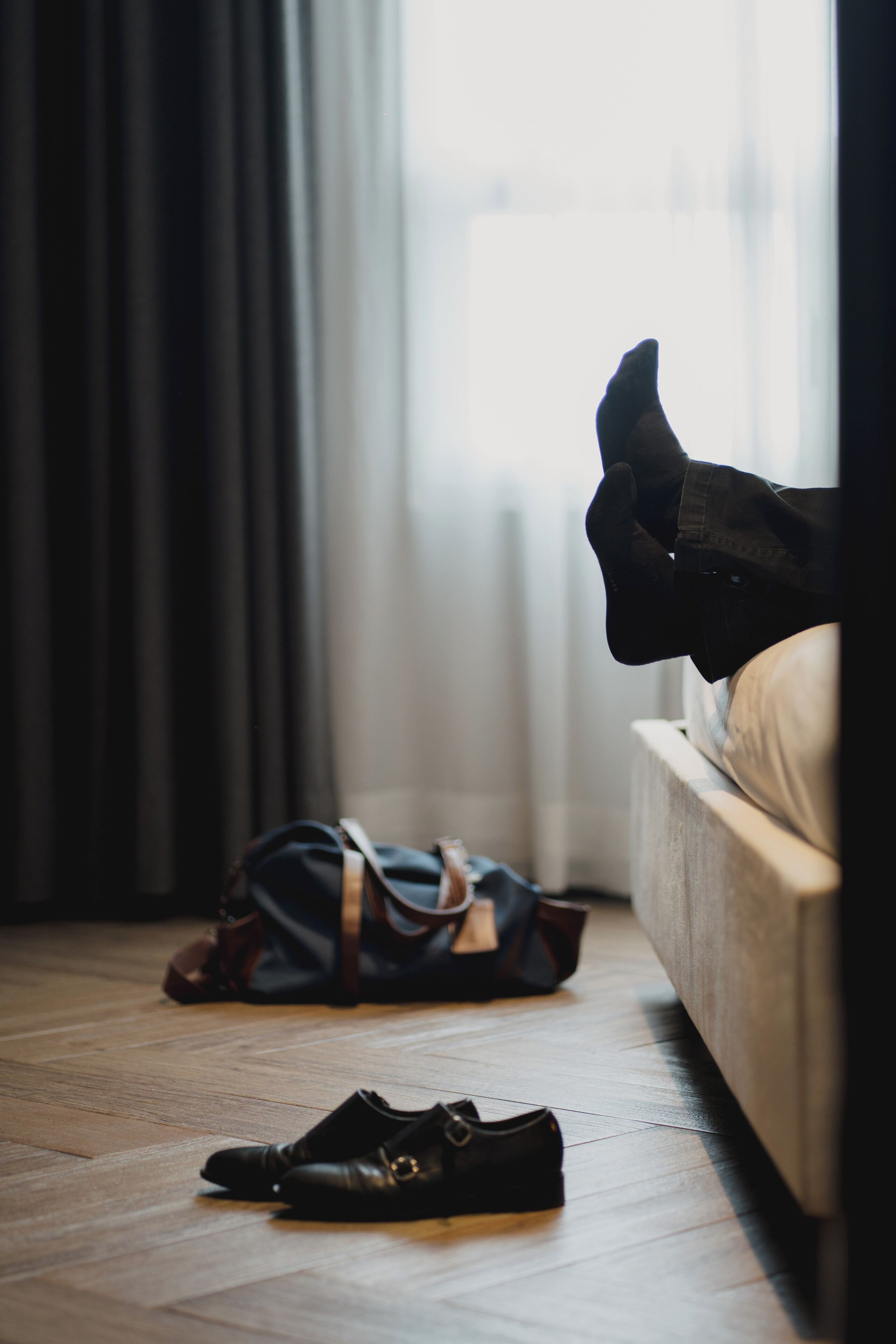 A room with a wooden floor, a bed, a black dog sitting on the bed, and a black shoe and a black bag on the floor.