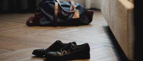 A room with a wooden floor, a bed, a black dog sitting on the bed, and a black shoe and a black bag on the floor.