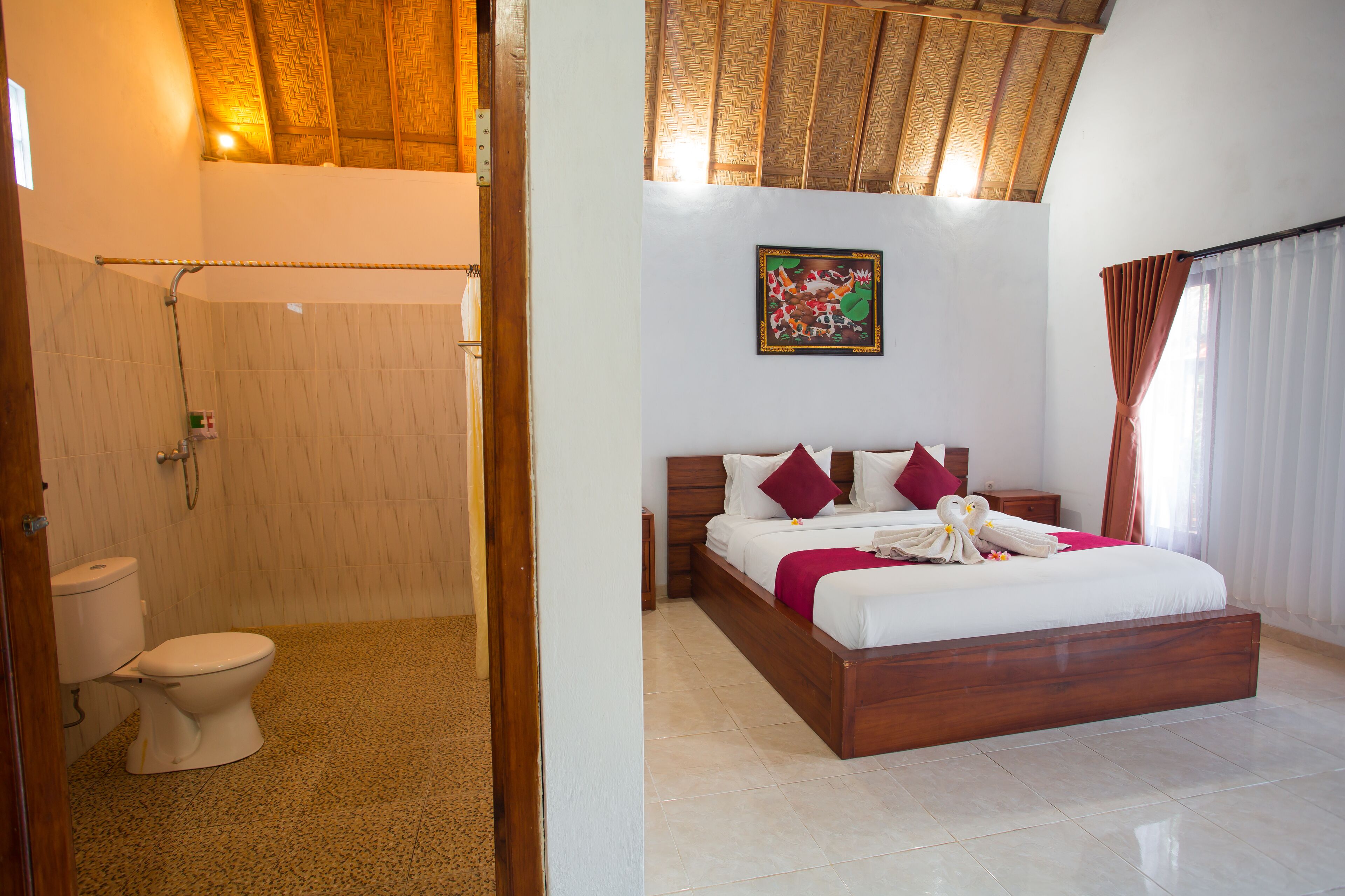 A bathroom with a toilet and shower, and a bedroom with a bed, pillows, and a painting on the wall.