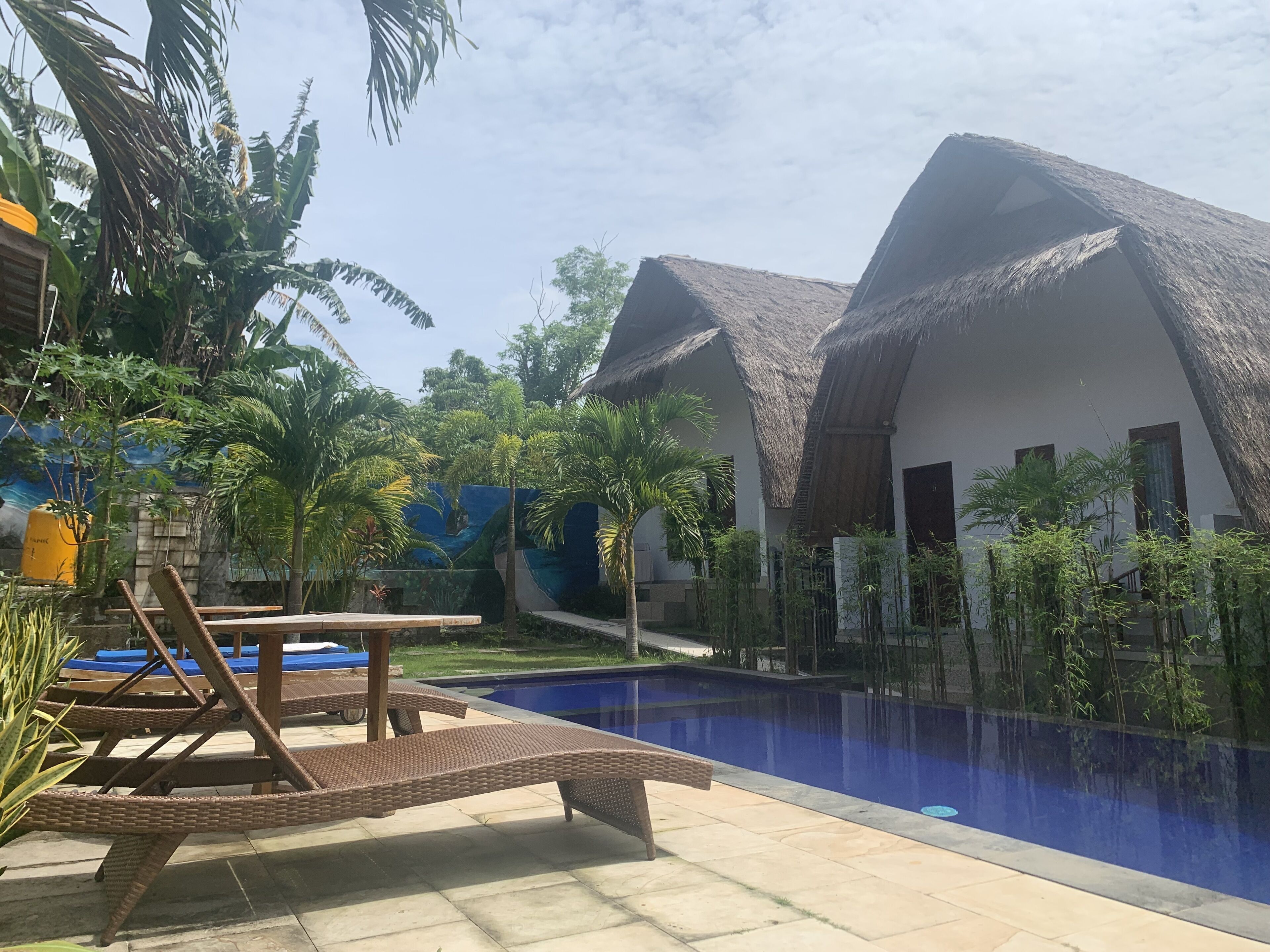 Un área junto a la piscina con sillones de descanso, un edificio con techo de paja y vegetación tropical.