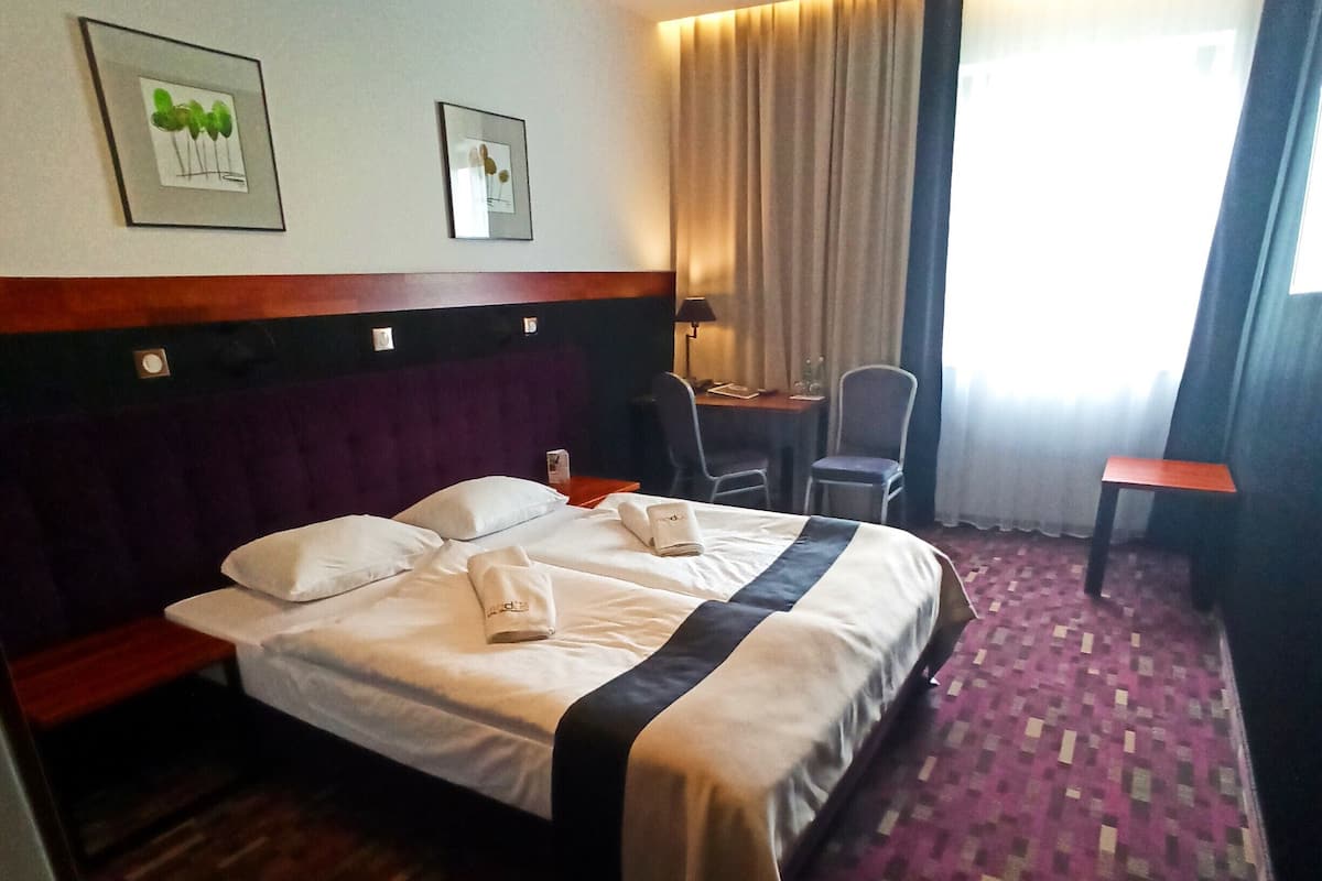 A hotel room with a bed, a desk, a chair, a small table, and a window with curtains.