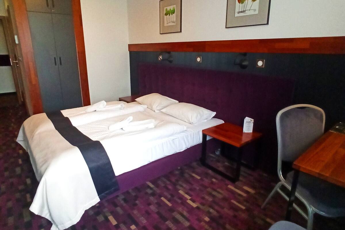 A hotel room with a bed, a desk, a chair, a wardrobe, and two framed pictures on the wall.