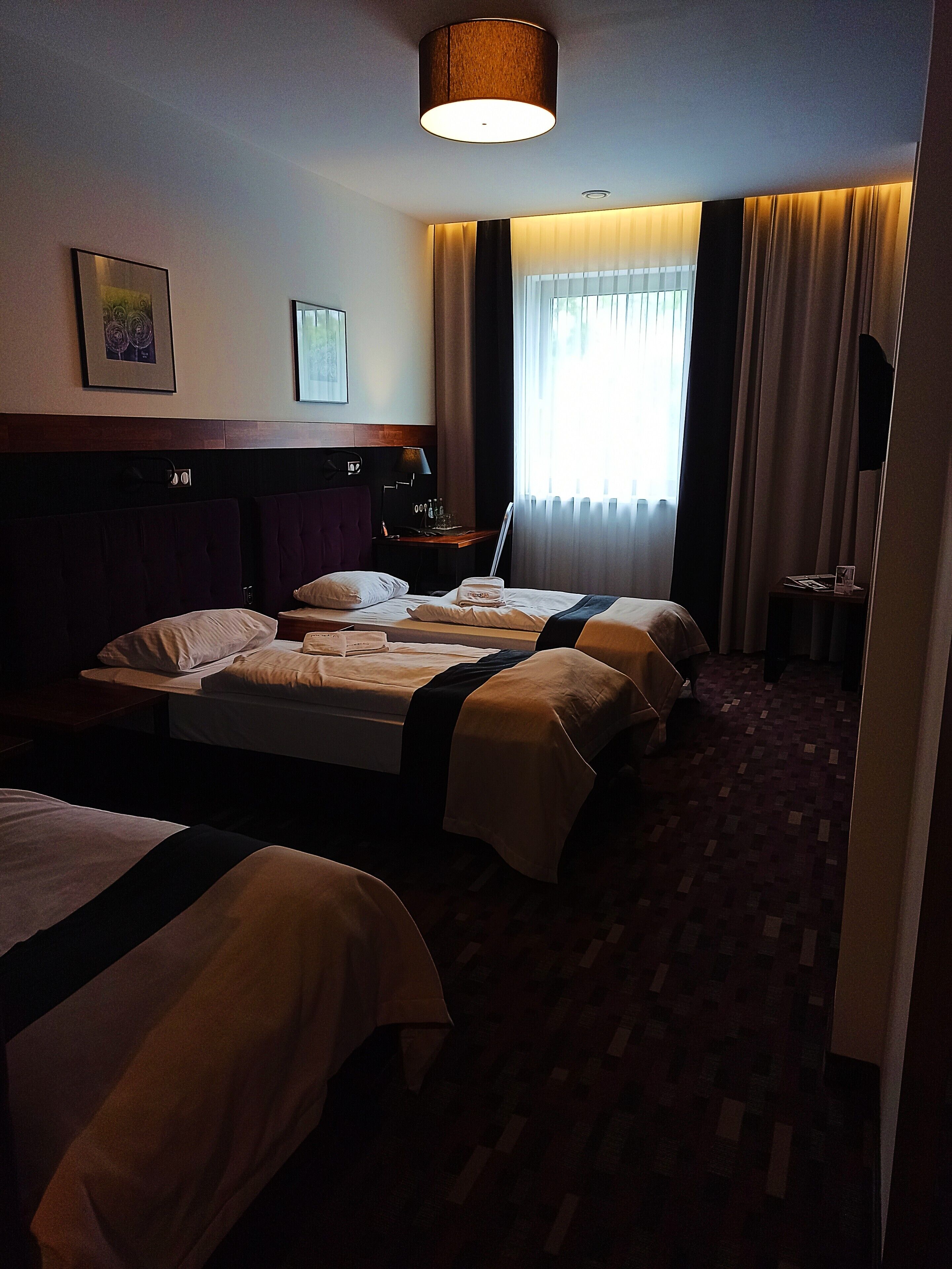 Hotellihuoneessa on suuri sänky, pienempi sänky, työpöytä, jossa on lamppu, ikkuna, jossa on verhot, ja seinällä taulu.