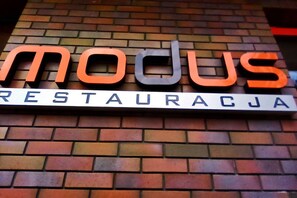Un letrero con la palabra 'modus' en letras grandes y 'RESTAURACJA' debajo, sujeto a una pared de ladrillos.