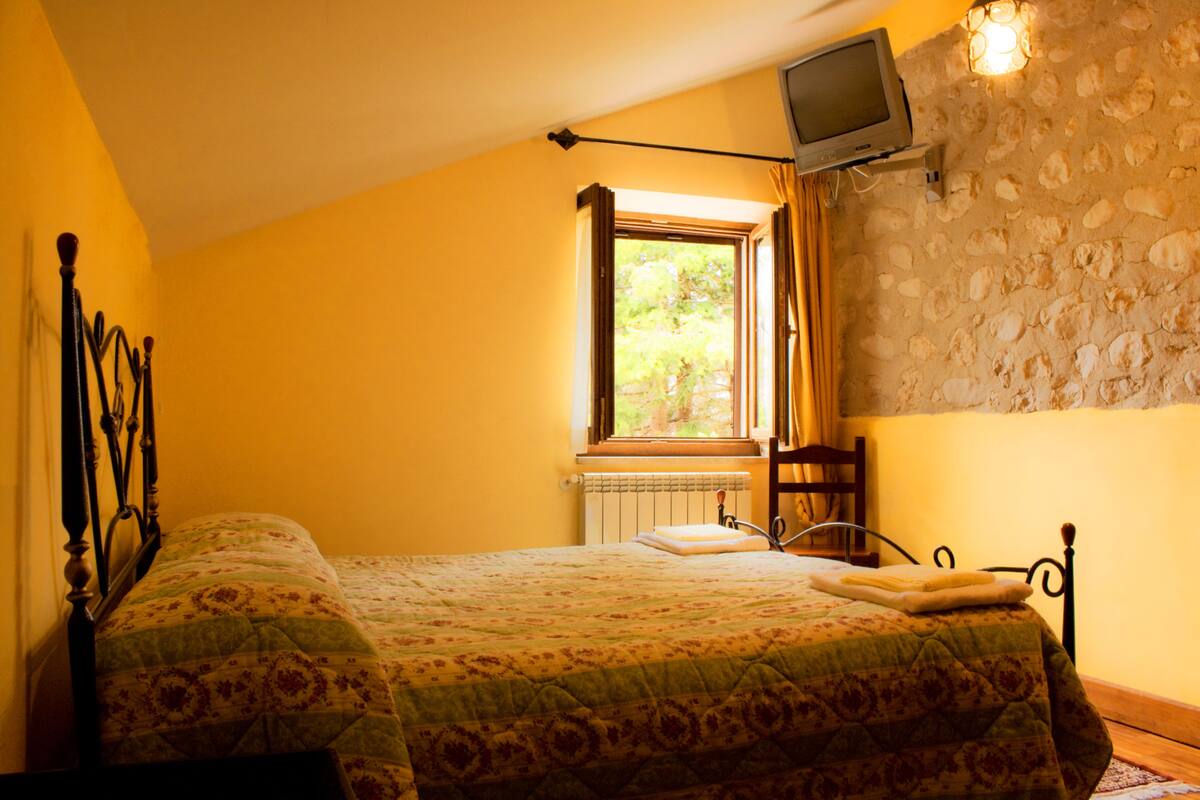 A bedroom with two beds, a stone wall, a window with curtains, and a television mounted on the wall.